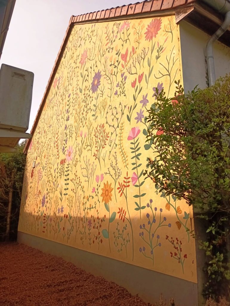 Muurschildering op gevel met fijne bloemen, planten en speelse botanische illustraties in zachte kleuren op een warme gele achtergrond, handgeschilderd en decoratief.
