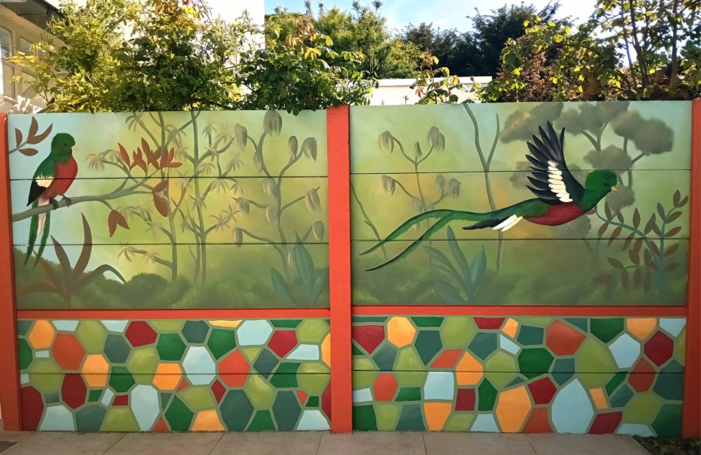Muurschildering met tropische quetzalvogels, jungleplanten en kleurrijk mozaïekpatroon op tuinmuur, handgeschilderd in groene en warme natuurtinten.
